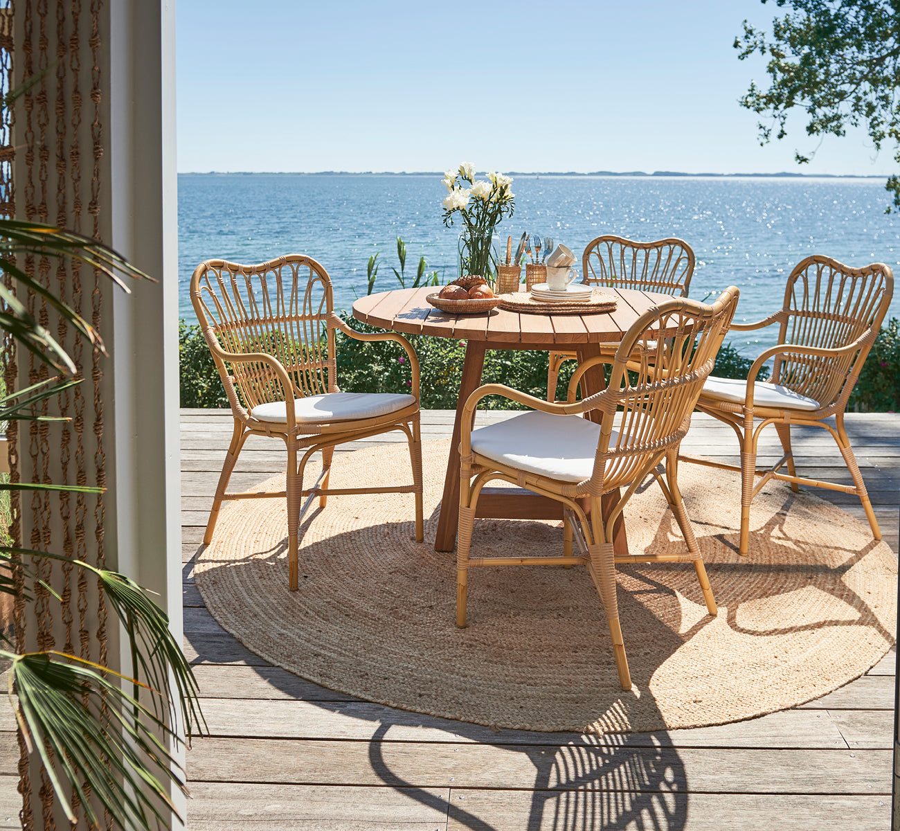 Dette er et livsstilsbillede af Margret Udendørs spisestuelænestol i natural aluminium artfibre. Dæk ved havet med rundt teakbord og havudsigt.