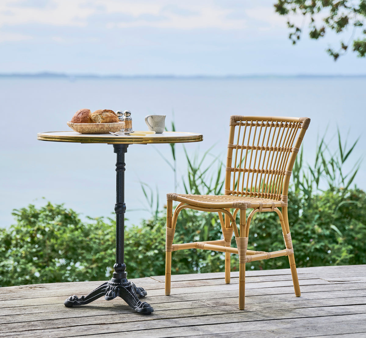 Dette er et livsstilsbillede af Elisabeth Udendørs Spisebordsstol i natural Aluminium ArtFibre, på et trædæk med et bistro-morgenbord med havudsigt.