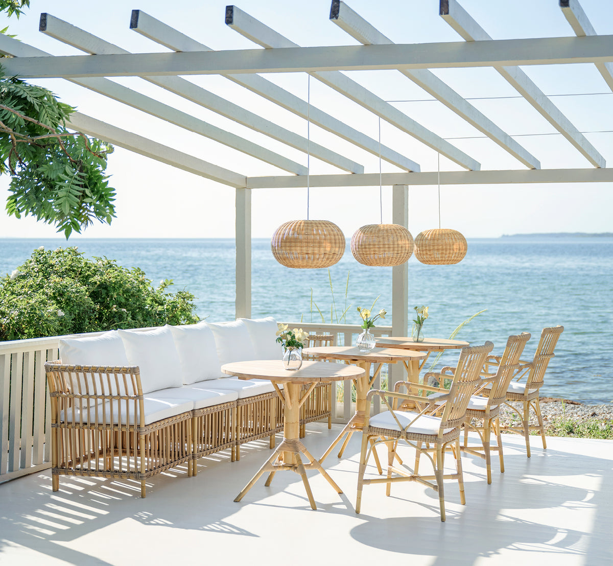 Dette er et livsstilsbillede af Piano Exterior Dining Armchair i natural aluminium artfibre. Under en pergola ved havet med rattanpendler.