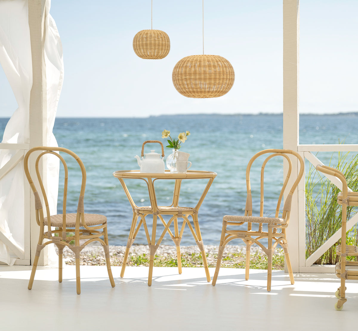 Dette er et livsstilsbillede af Lulu Udendørs spisebordsstol i natural aluminium artfibre. På en strandterrasse med et rundt bord og havudsigt.