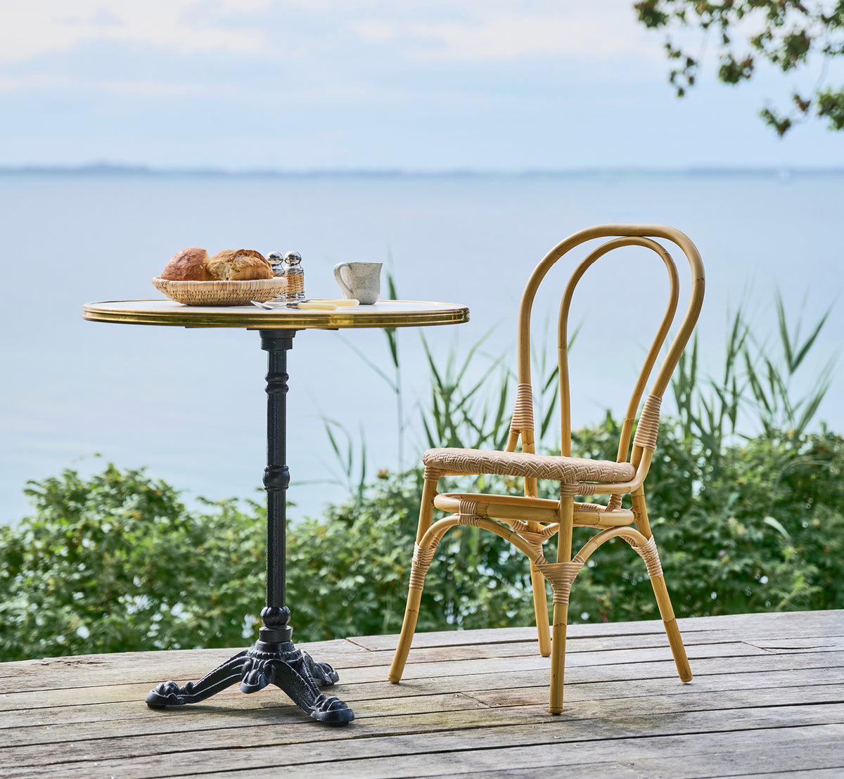 Dette er et livsstilsbillede af Lulu Udendørs spisebordsstol i natural aluminium artfibre. Ved et havnefront-bistrobord med bagværk og roligt hav.