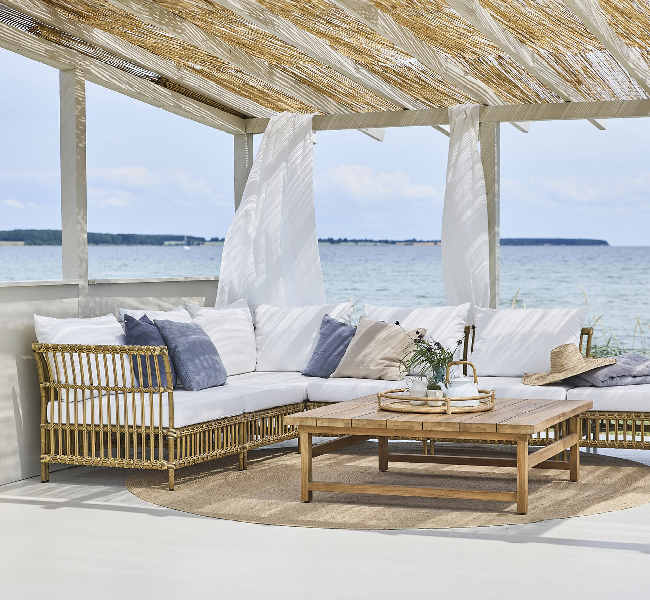 Dette er et livsstilsbillede af Maggie Sofa Hjørnemodul i antique aluminium artfibre. Strandpavillon med sofasæt og havudsigt.