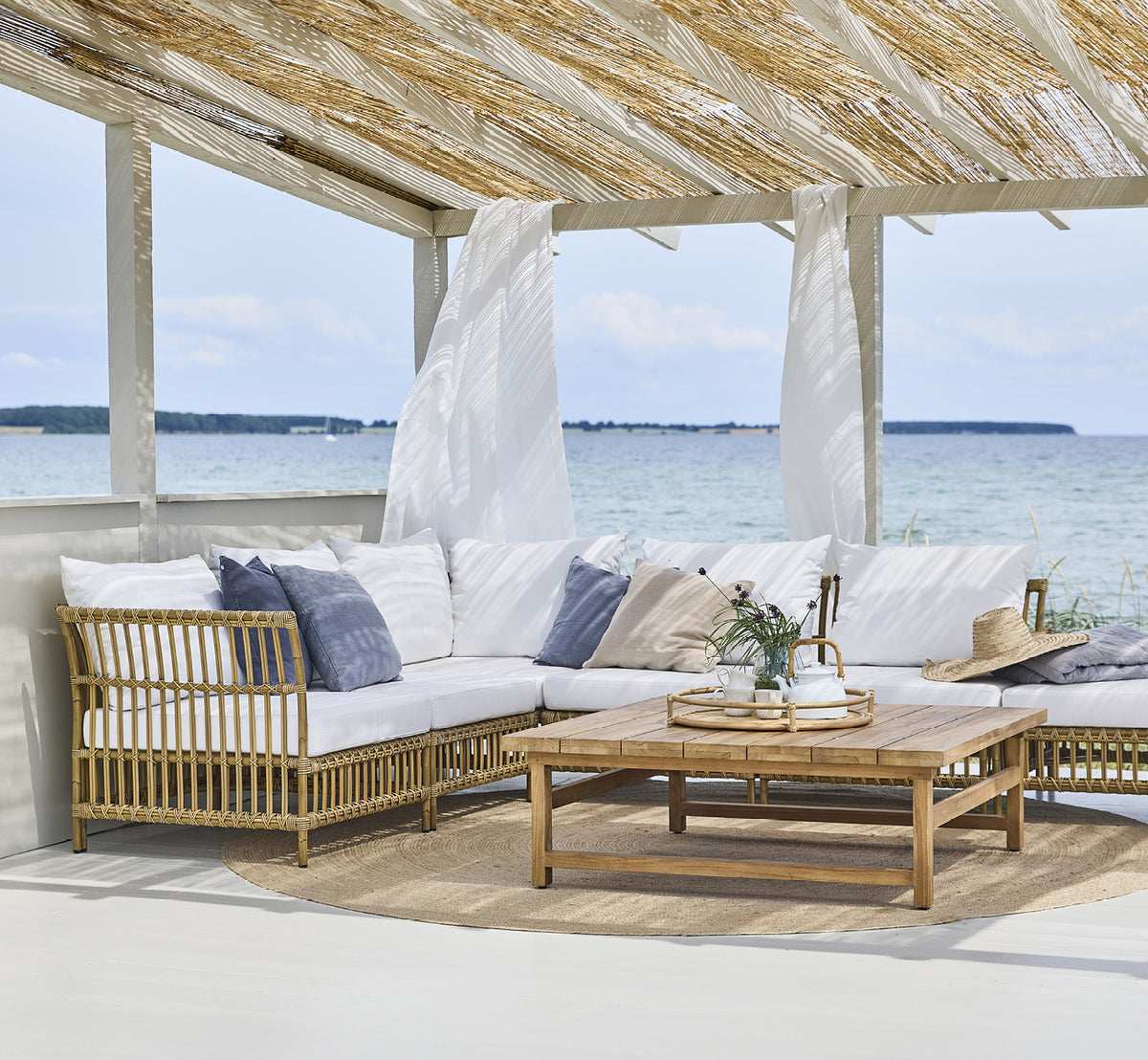 Dette er et livsstilsbillede af Maggie Sofa Hjørnemodul i antique aluminium artfibre. Strandpavillon med sofasæt og havudsigt.