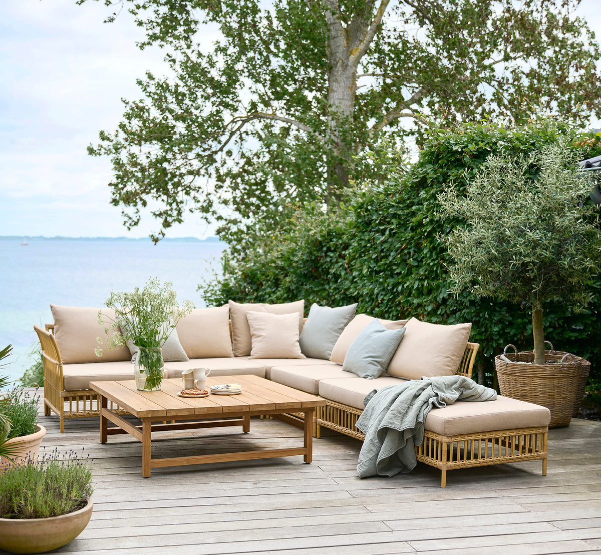 Dette er et livsstilsbillede af Maggie Sofa Hjørnemodul i natural aluminium artfibre. Trædæk ved havet med puder og havudsigt.