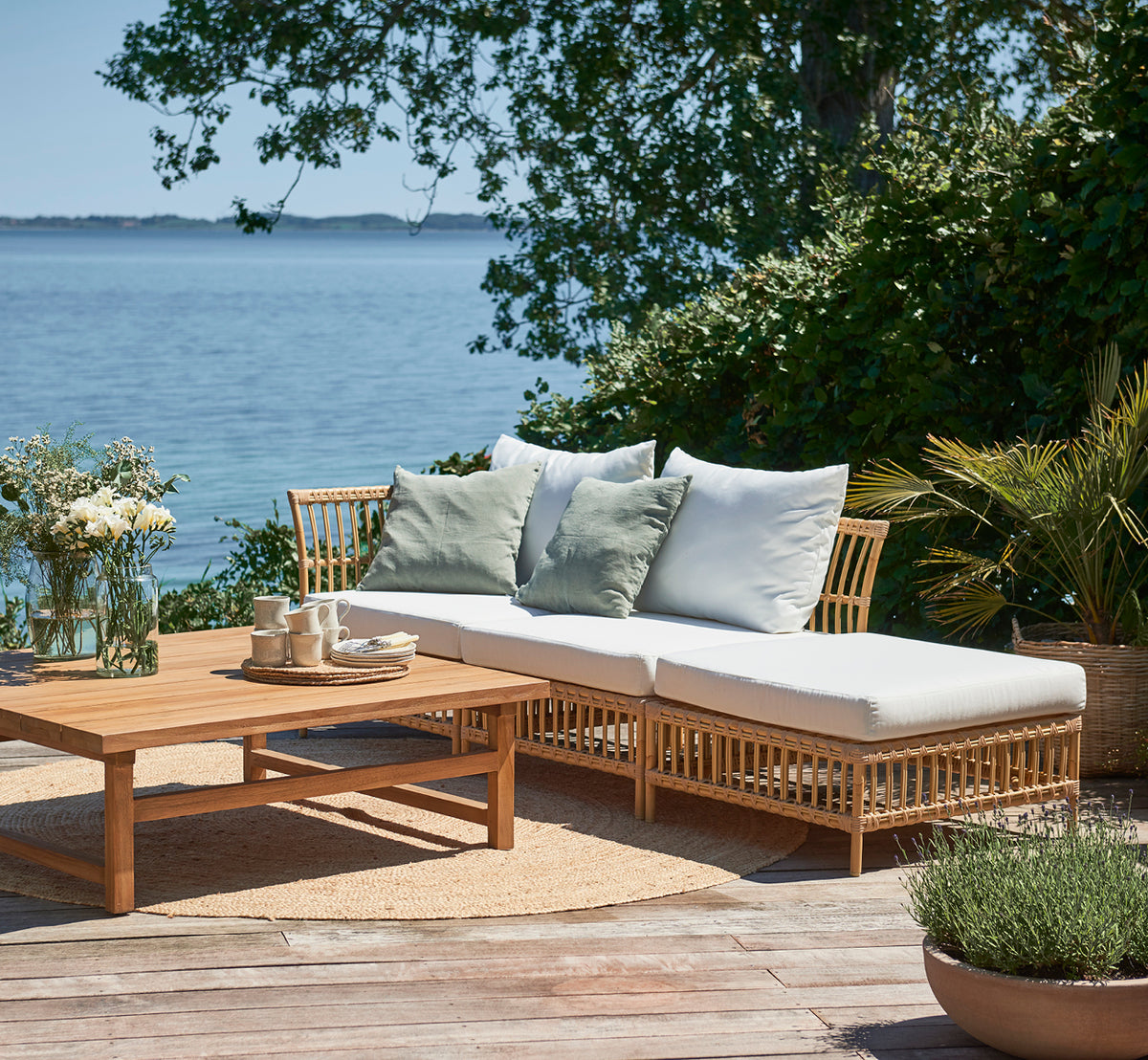 Dette er et livsstilsbillede af Maggie Sofa Centermodul i natural aluminium artfibre. Dæk ved havet med puder, planter og havudsigt.