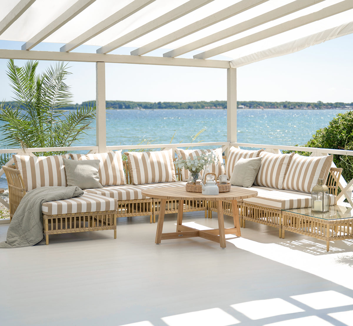 Dette er et livsstilsbillede af Maggie Sofa Centermodul i natural aluminium artfibre. Overdækket terrasse med stribede puder og havbaggrund.