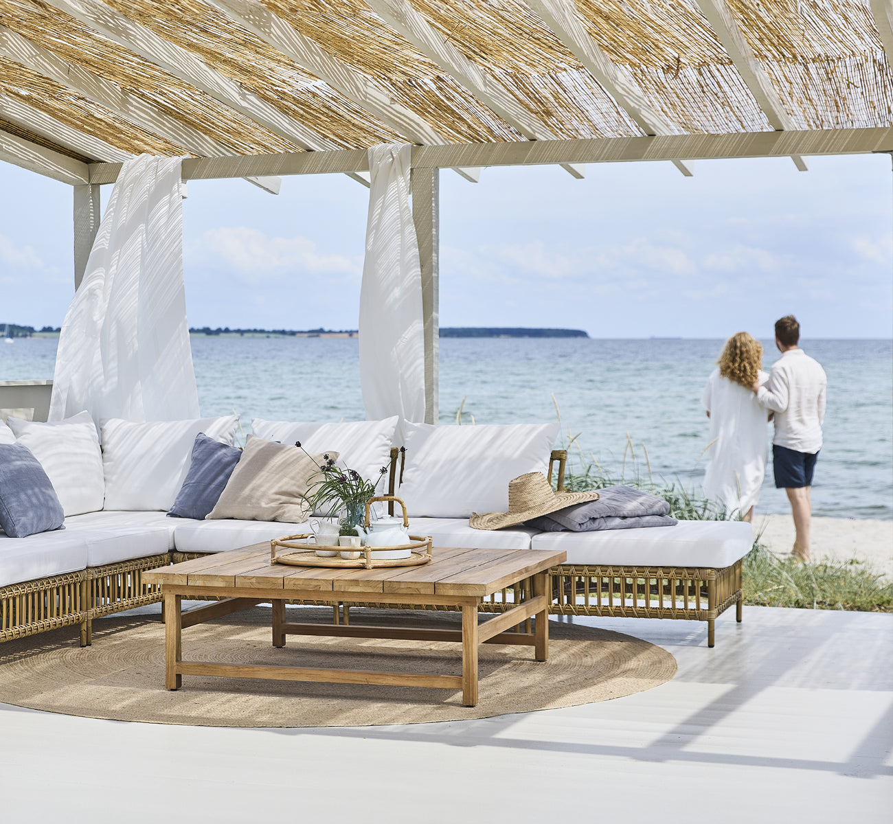 Dette er et livsstilsbillede af Maggie Sofa Puf Modul m. Glas i antique aluminium artfibre. Strandpavillon med par og havudsigt.
