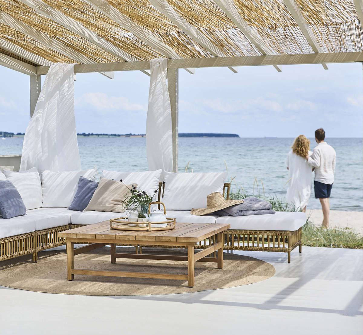 Dette er et livsstilsbillede af Maggie Sofa Puf Modul m. Glas i antique aluminium artfibre. Strandpavillon med par og havudsigt.