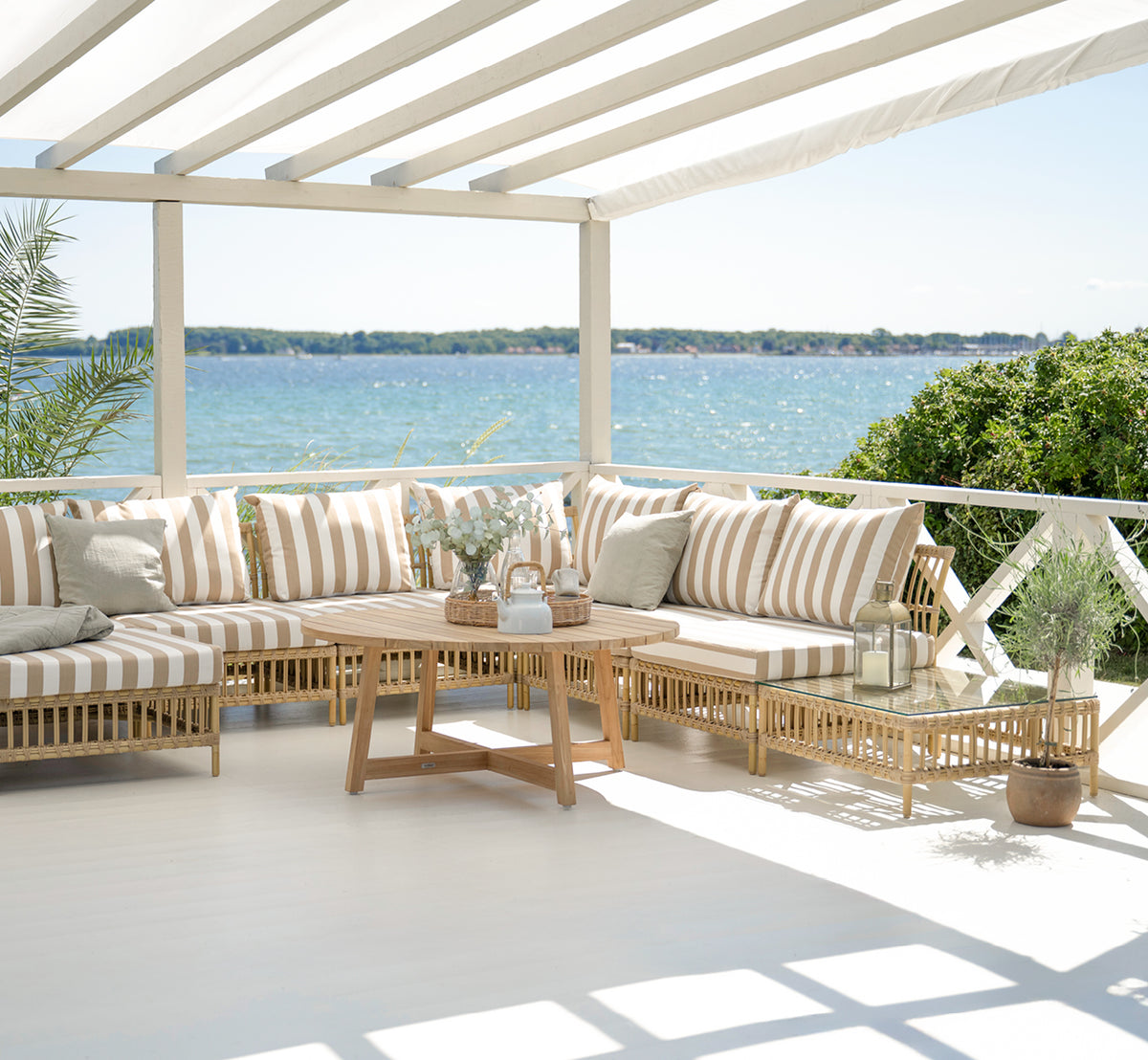 Dette er et livsstilsbillede af Maggie Sofa Puf Modul m. Glas i natural aluminium artfibre. Overdækket terrasse med stribede puder og havudsigt.
