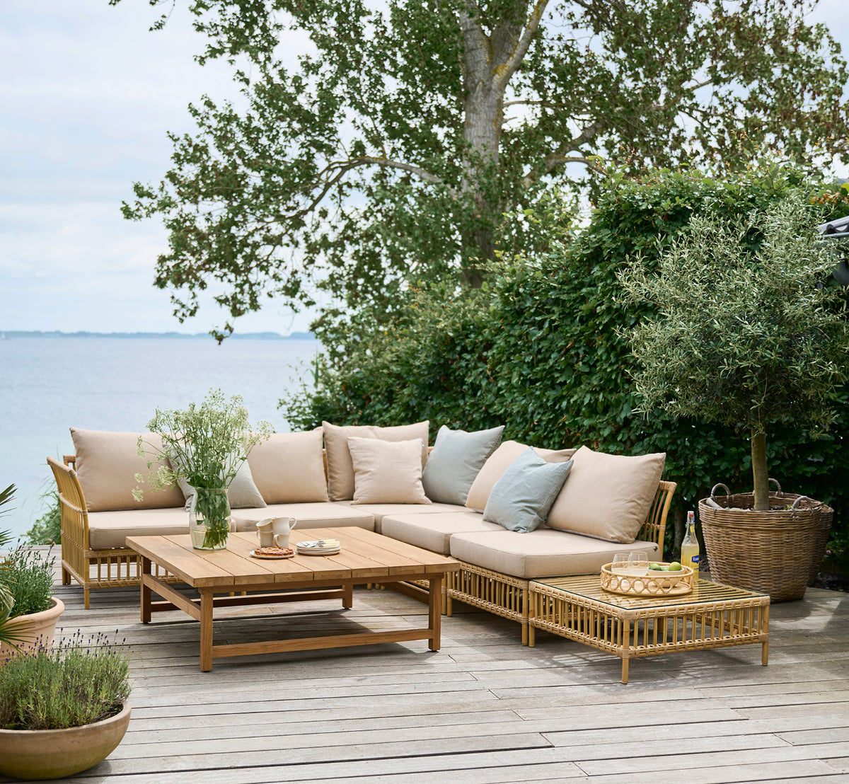 Dette er et livsstilsbillede af Maggie Sofa Puf Modul m. Glas i natural aluminium artfibre. Dæk ved havet med sofasæt og grønt omkring.