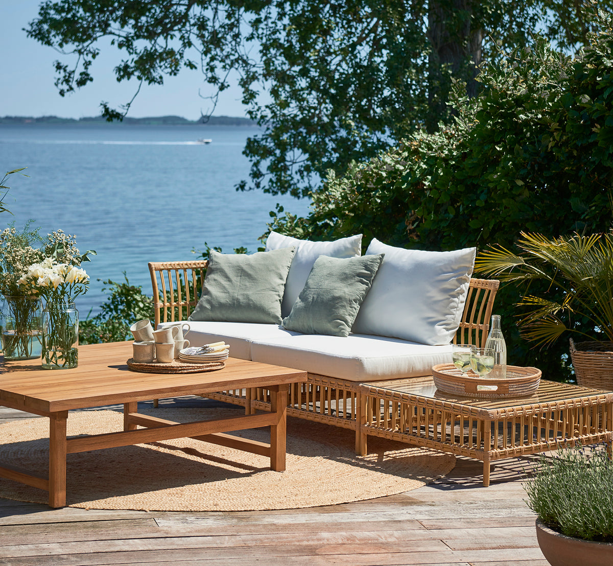 Dette er et livsstilsbillede af Maggie Sofa Puf Modul m. Glas i natural aluminium artfibre. Dæk ved havet med sofabord og grønne puder.