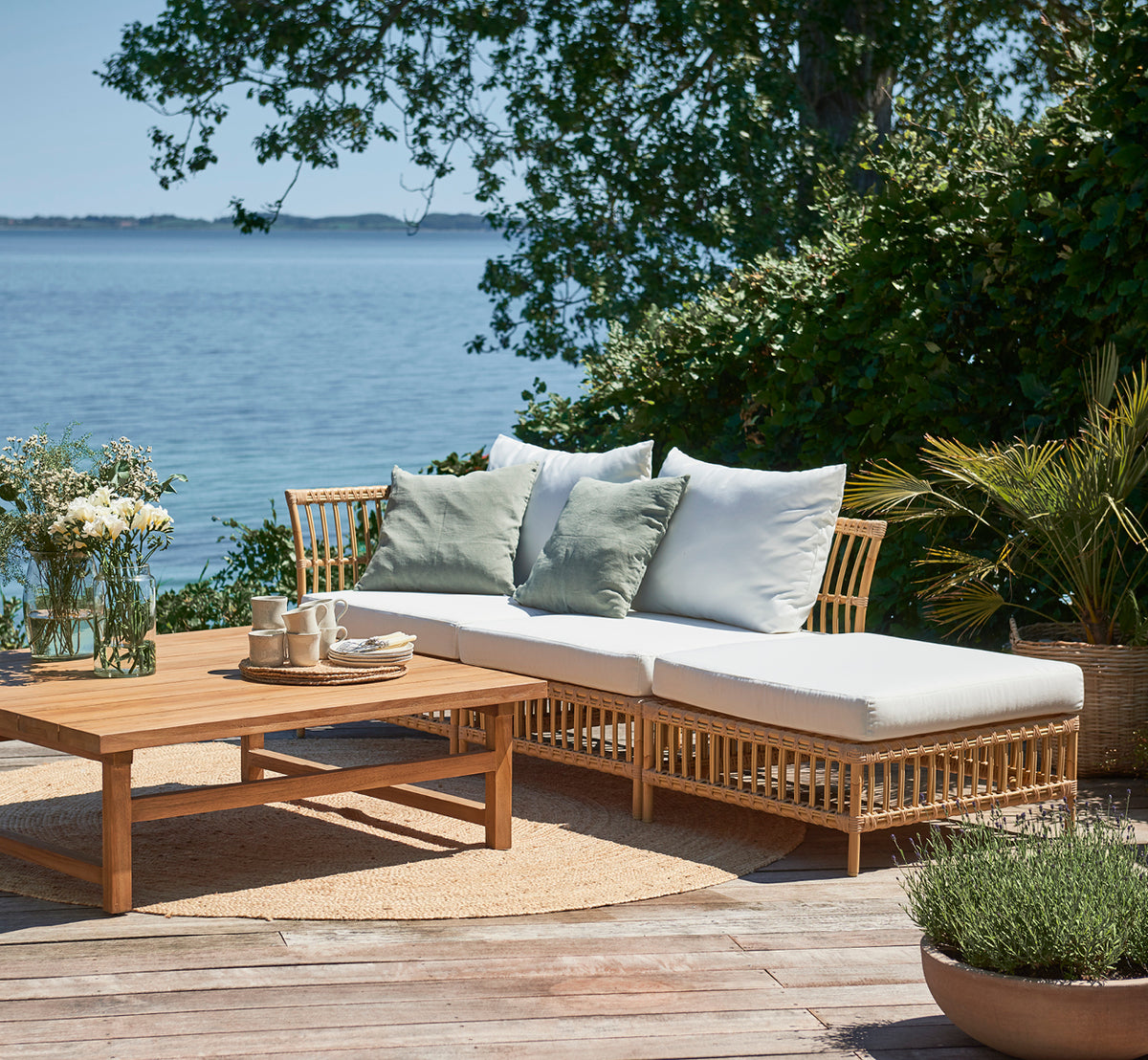 Dette er et livsstilsbillede af Maggie Sofa Puf Modul m. Glas i natural aluminium artfibre. Dæk ved havet med sofabord og grønne puder.