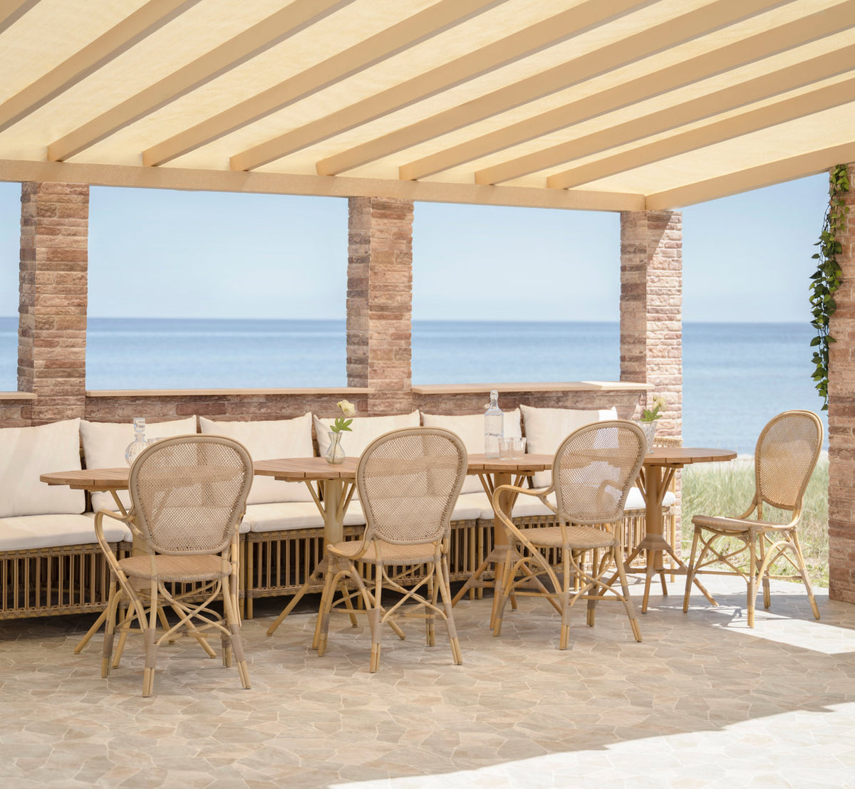 Dette er et livsstilsbillede af Meghan Udendørs spisebænk-Corner Module i natural aluminium artfibre. Under en pergola ved havet.