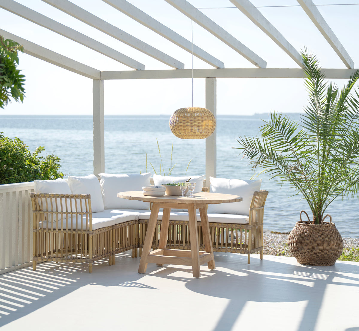 Dette er et livsstilsbillede af Meghan Exterior Dining Bench Center Module i natural aluminium artfibre. Omkring et rundt bord ved havet.