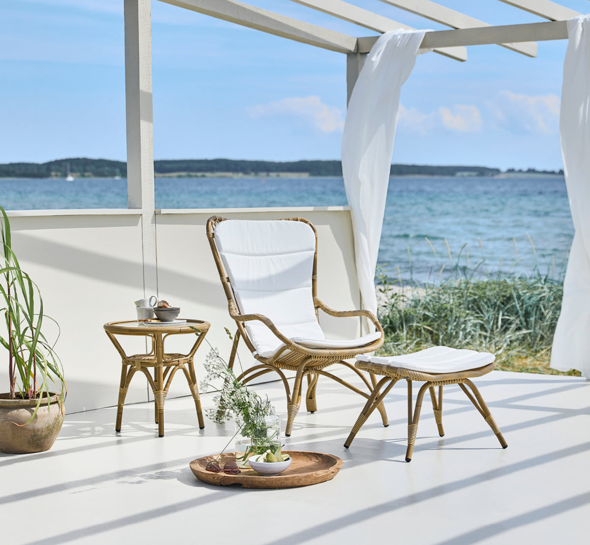 Dette er et livsstilsbillede af Monet Exterior Lounge Chair i antique aluminium artfibre. Med fodskammel på et dæk i en strandpavillon.