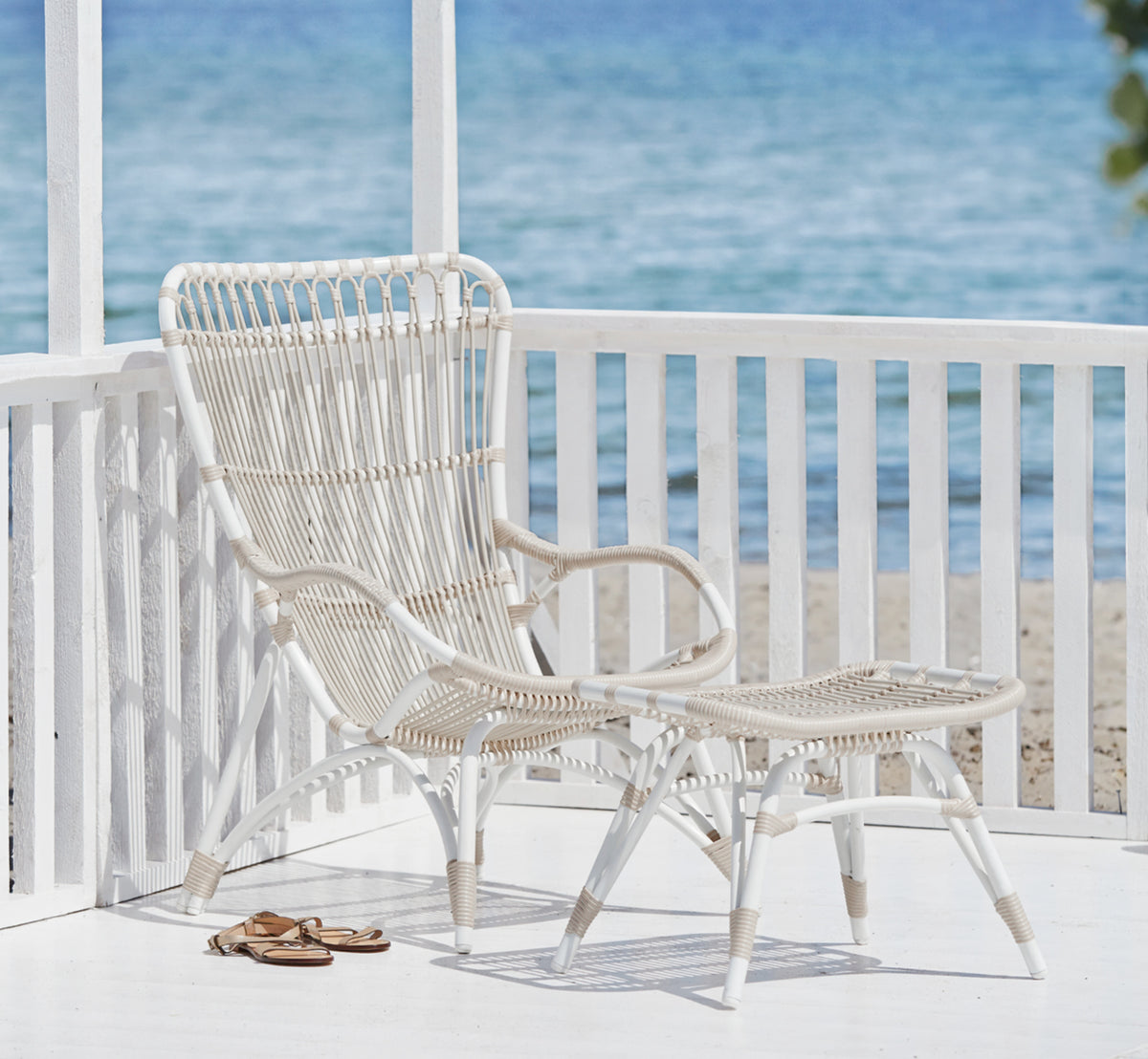 Dette er et livsstilsbillede af Monet Exterior Lounge Chair i dove white aluminium artfibre. Med fodskammel på en veranda ved havet.