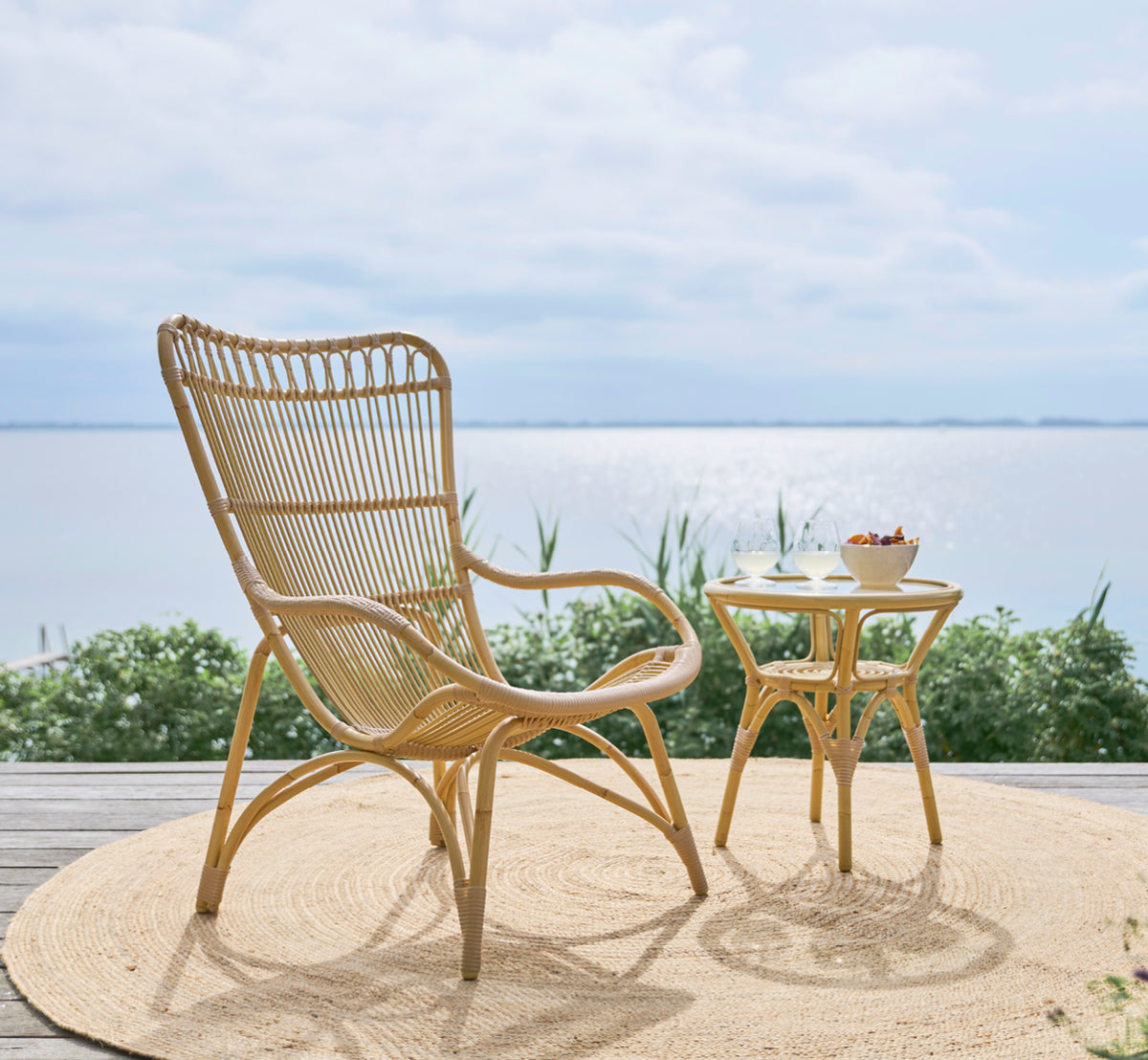 Dette er et livsstilsbillede af Monet Exterior Lounge Chair i natural aluminium artfibre. På et trædæk med jutetæppe og havudsigt.