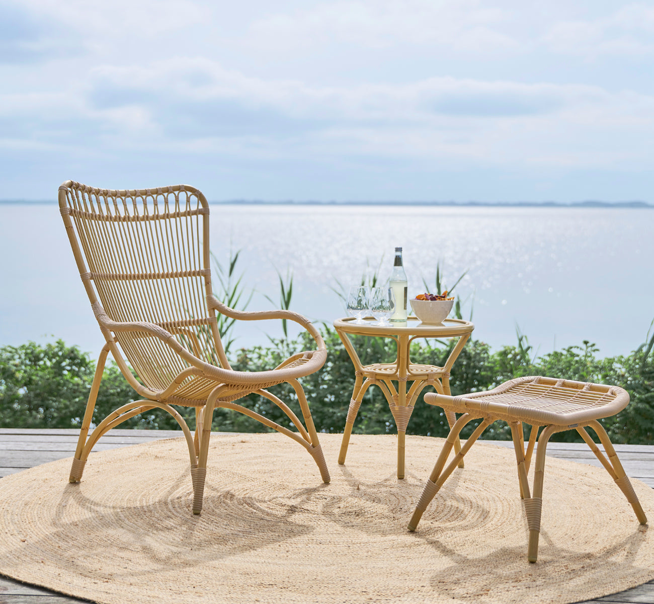 Dette er et livsstilsbillede af Monet Exterior Lounge Chair i natural aluminium artfibre. Med fodskammel på et dæk med udsigt over havet.