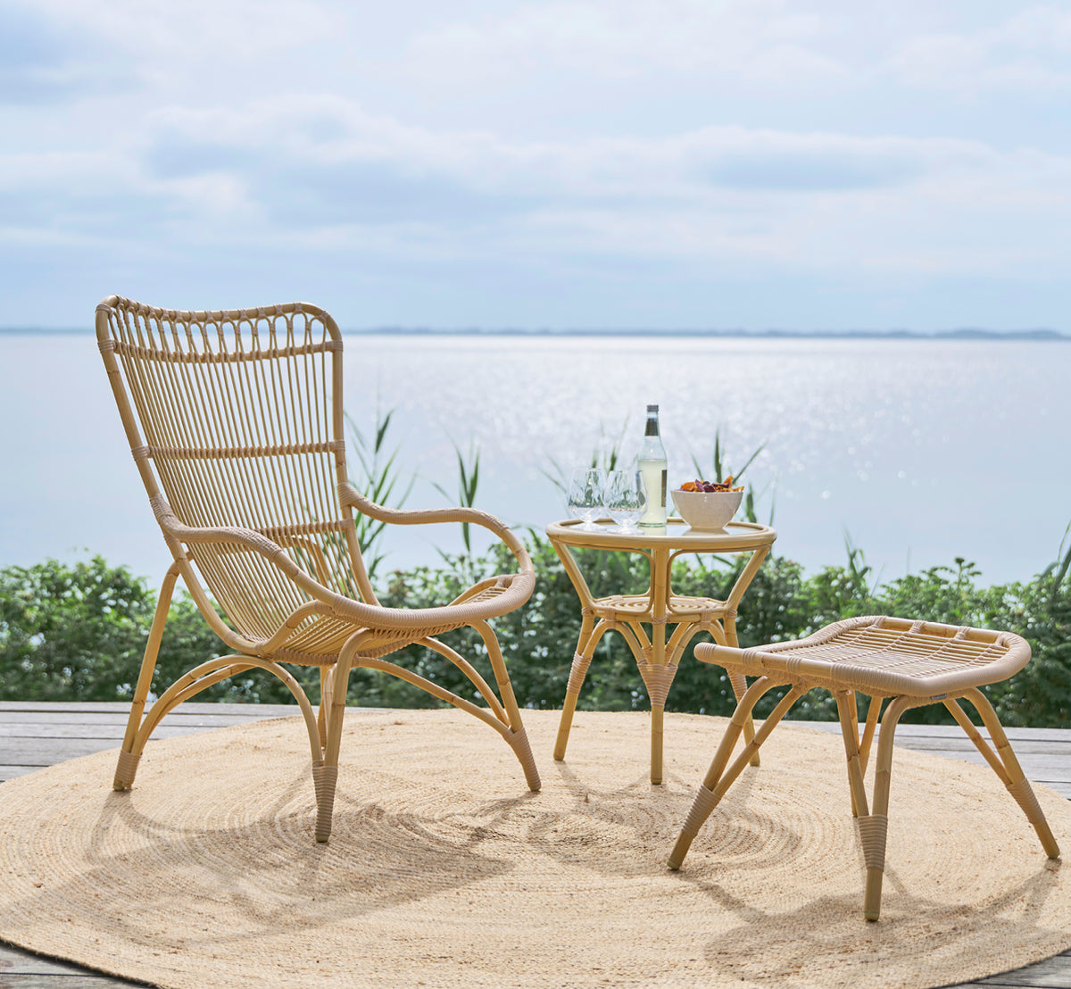 Dette er et livsstilsbillede af Monet Exterior Lounge Chair i natural aluminium artfibre. Med fodskammel på et dæk med udsigt over havet.