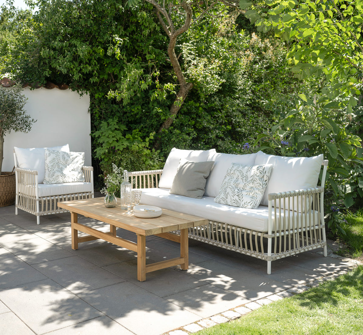 Dette er et livsstilsbillede af Caroline Udendørs sofa i dove White Aluminium ArtFibre, på et trædæk ved vandet med kystnær vegetation og havudsigt.