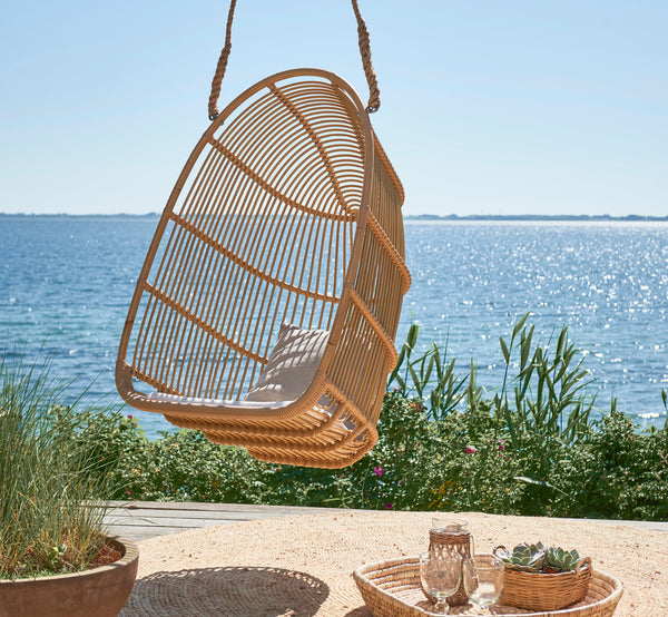 Dette er et livsstilsbillede af Renoir Udendørs hængestol i natural aluminium artfibre. Hængende på en strandterrasse med bakke og sukkulenter nedenfor.