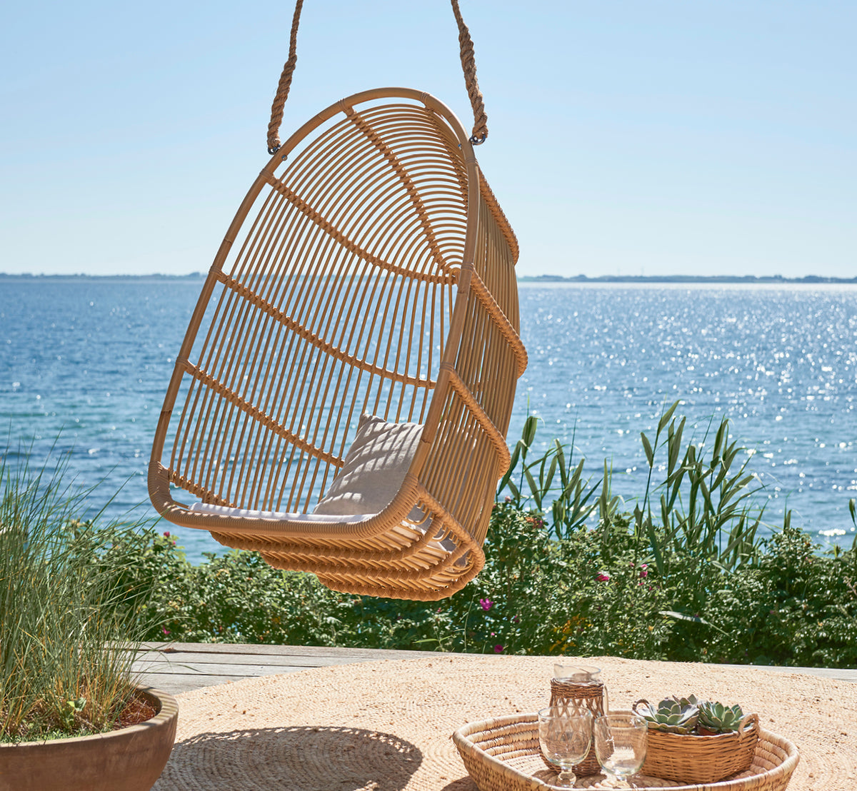 Dette er et livsstilsbillede af Renoir Udendørs hængestol i natural aluminium artfibre. Hængende på en strandterrasse med bakke og sukkulenter nedenfor.