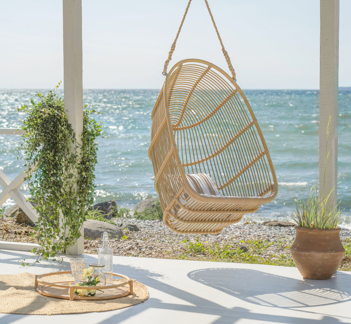 Dette er et livsstilsbillede af Renoir Udendørs hængestol i natural aluminium artfibre. Hængende på en strandterrasse med bakke og sukkulenter nedenfor.