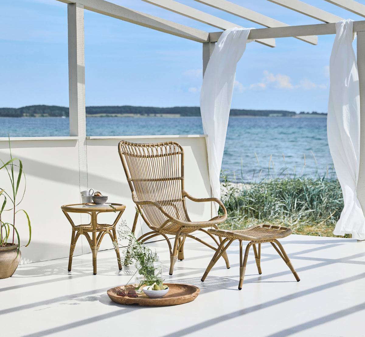 Dette er et livsstilsbillede af Tony Exterior Side Table i antique aluminium artfibre. Ved siden af en lænestol i en strandpavillon.