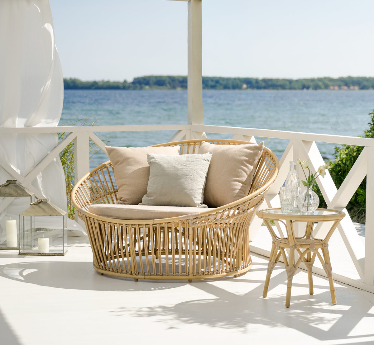 Dette er et livsstilsbillede af Tony Exterior Side Table i natural aluminium artfibre. På et trædæk med jutetæppe ved havet.