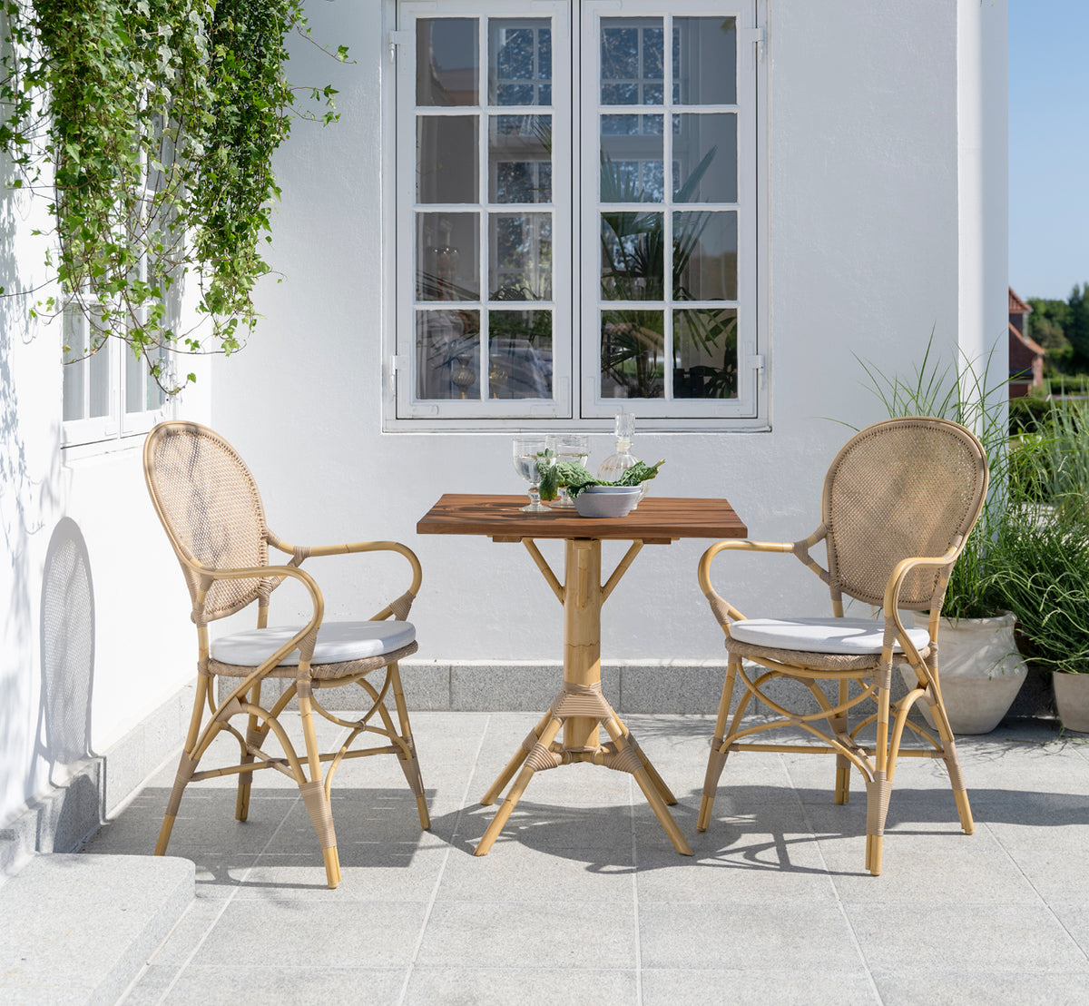 Dette er et livsstilsbillede af Nicole Cafe Table i natural aluminium artfibre. På en stenterrasse med Ofelia-stole og grønne planter.