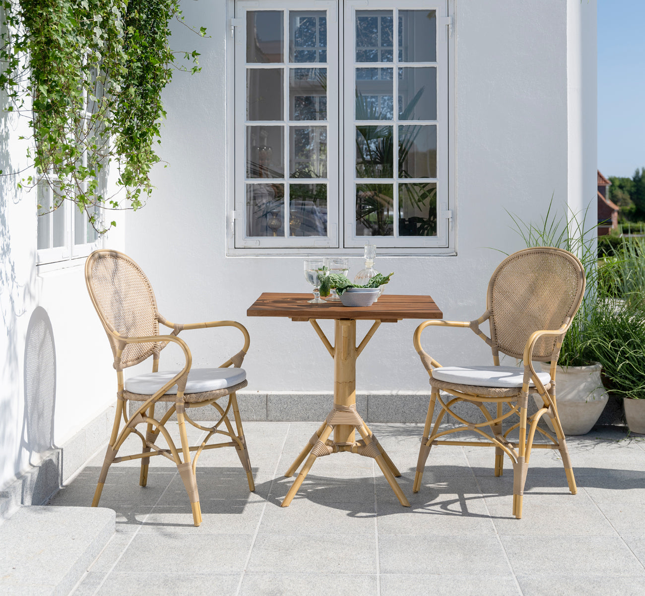 Dette er et livsstilsbillede af Nicole Cafe Table i natural aluminium artfibre. På en stenterrasse med Ofelia-stole og grønne planter.