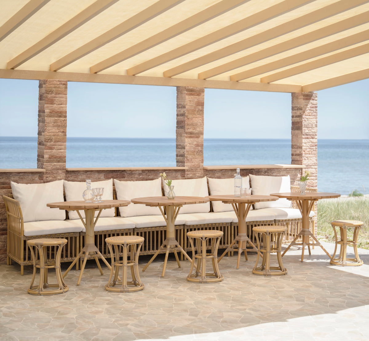 Dette er et livsstilsbillede af Nicole Cafébord-Teak Ø80 cm i natural aluminium artfibre. Bænk med skamler og cafébord under en pergola.