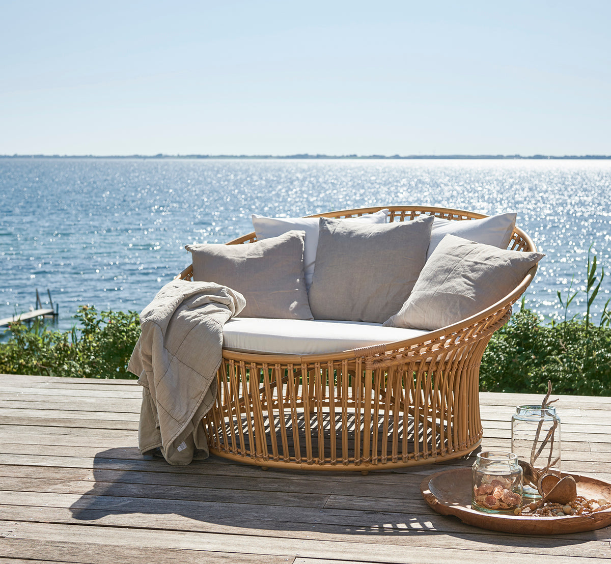 Dette er et livsstilsbillede af Olympia Nest Exterior i natural aluminium artfibre. I en strandpavillon med puder og havudsigt.