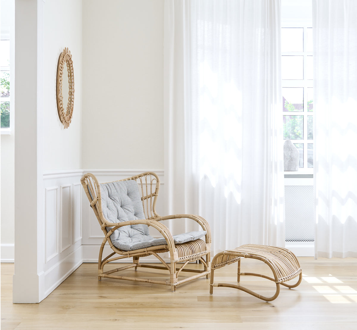 Dette er et livsstilsbillede af Teddy Lænestol i polished natural rattan. Placeret på et sildebensgulv ved et gardinvindue med lille spejl.