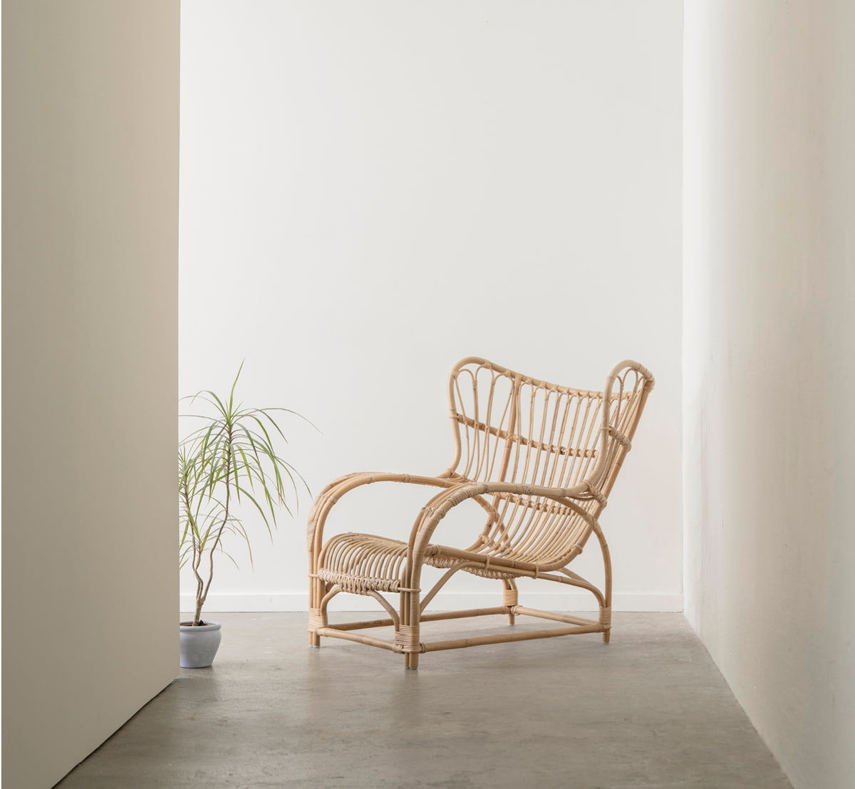 Dette er et livsstilsbillede af Teddy Lænestol i polished natural rattan. Placeret på et sildebensgulv ved et gardinvindue med lille spejl.