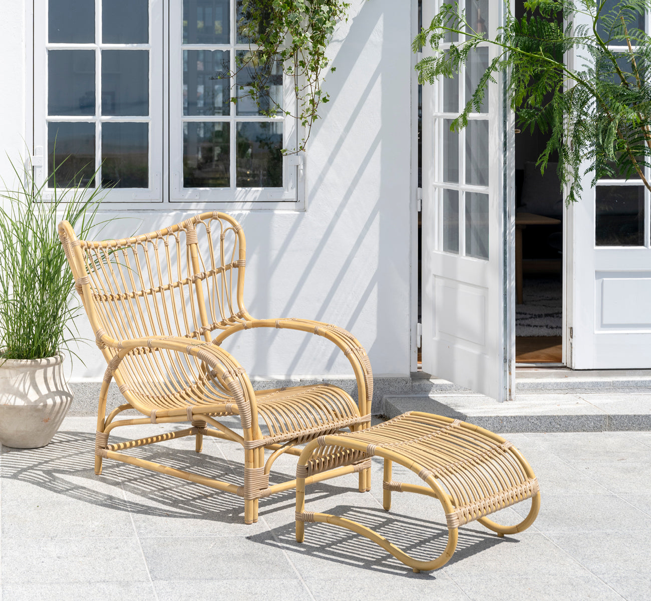Dette er et livsstilsbillede af Teddy Udendørs lænestol i natural aluminium artfibre. Placeret på en solbeskinnet brolagt terrasse ved en hvid-indrammet døråbning.