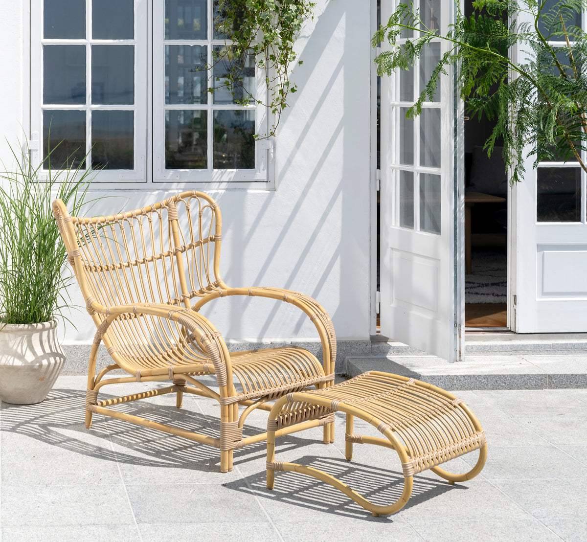 Dette er et livsstilsbillede af Teddy Udendørs lænestol i natural aluminium artfibre. Placeret på en solbeskinnet brolagt terrasse ved en hvid-indrammet døråbning.