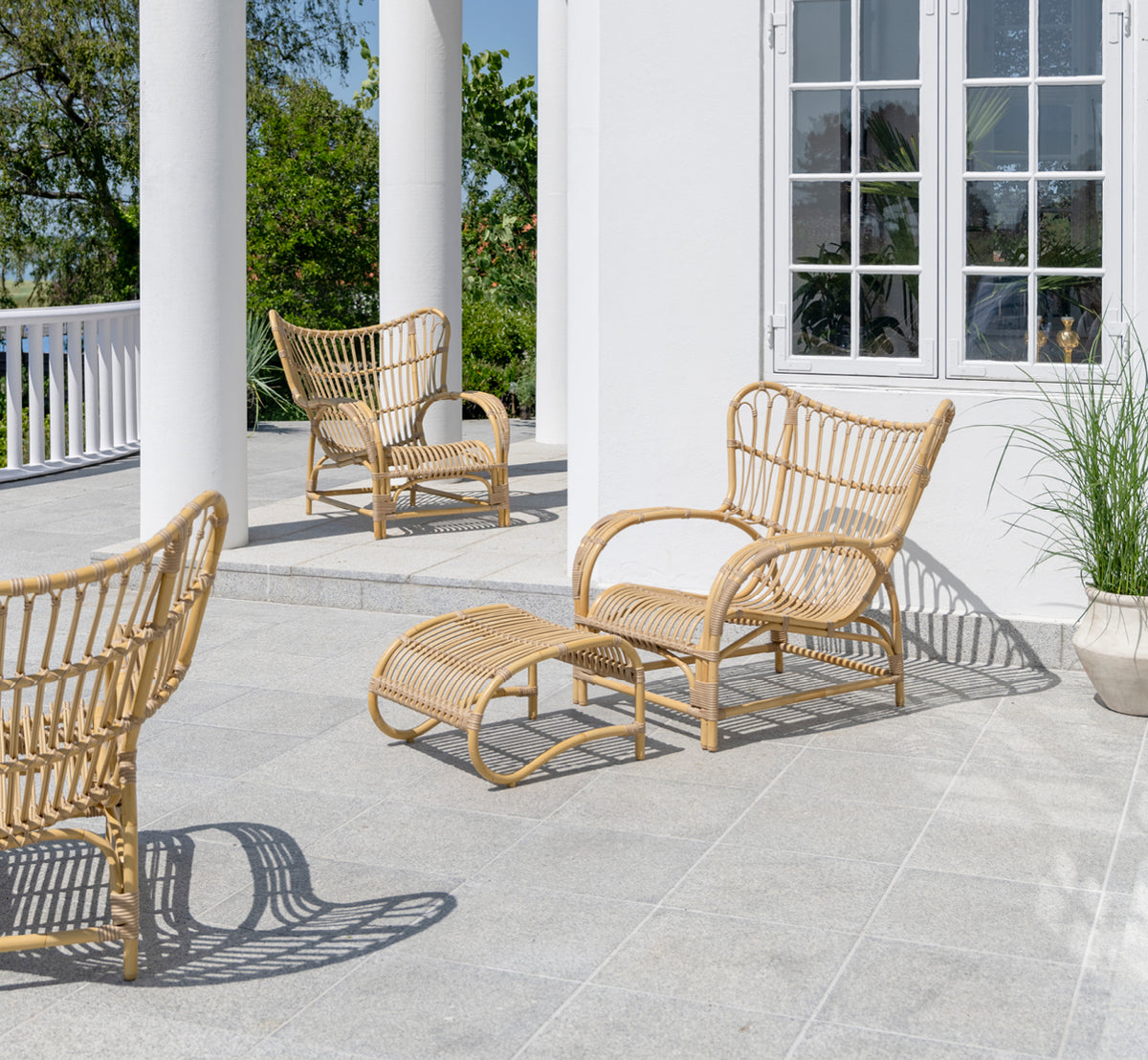 Dette er et livsstilsbillede af Teddy Udendørs lænestol i natural aluminium artfibre. Placeret på en solbeskinnet brolagt terrasse ved en hvid-indrammet døråbning.