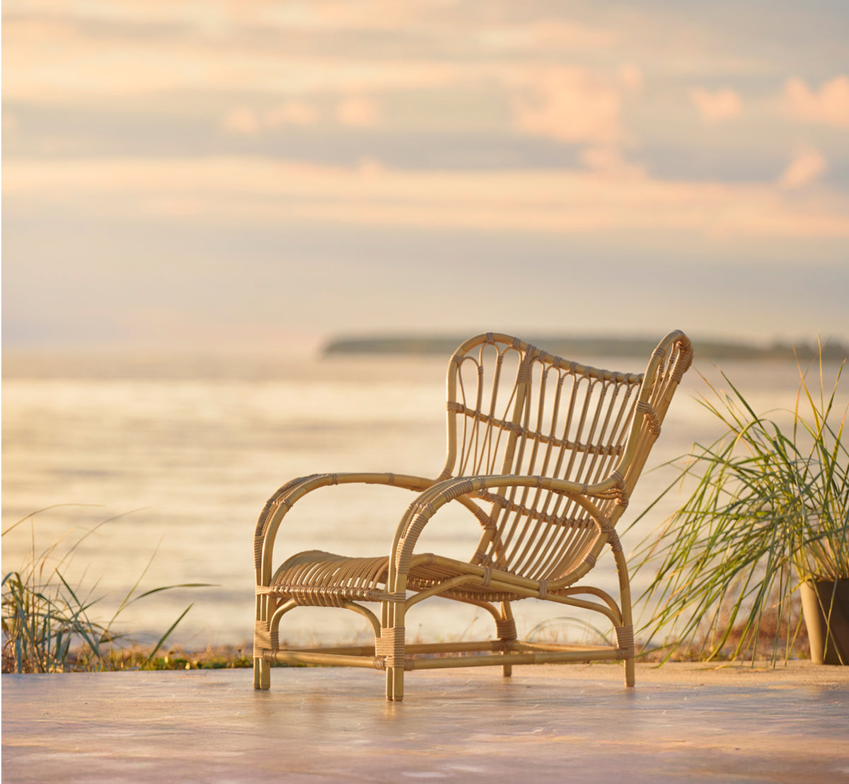 Dette er et livsstilsbillede af Teddy Udendørs lænestol i natural aluminium artfibre. Placeret på en solbeskinnet brolagt terrasse ved en hvid-indrammet døråbning.