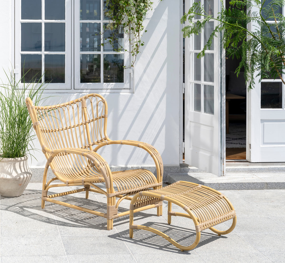 Dette er et livsstilsbillede af Teddy Udendørs fodskammel i natural aluminium artfibre. Kombineret med den matchende lænestol på en solbeskinnet brolagt terrasse.