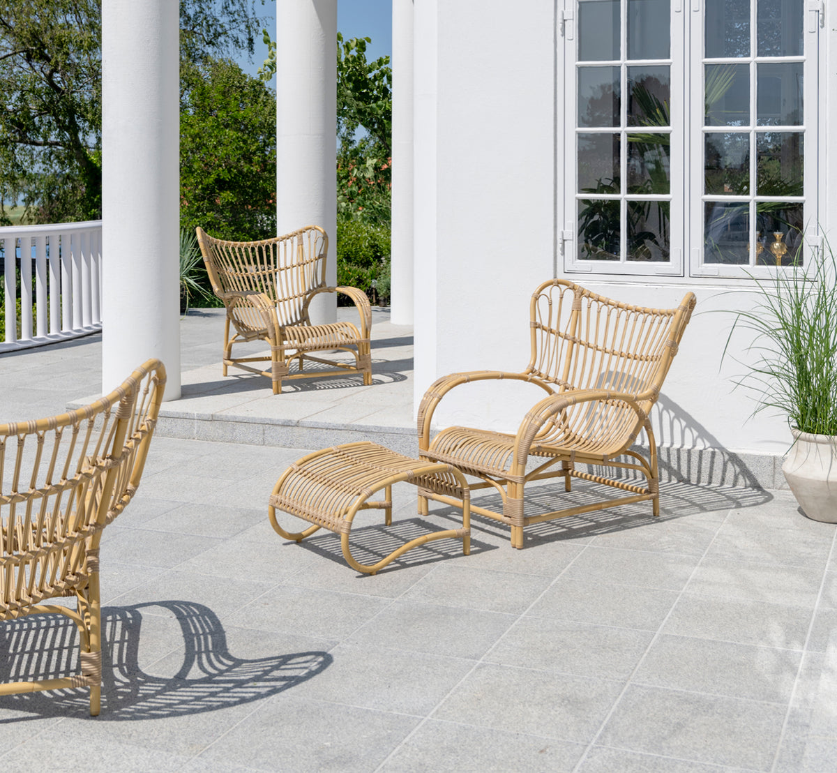Dette er et livsstilsbillede af Teddy Udendørs fodskammel i natural aluminium artfibre. Kombineret med den matchende lænestol på en solbeskinnet brolagt terrasse.