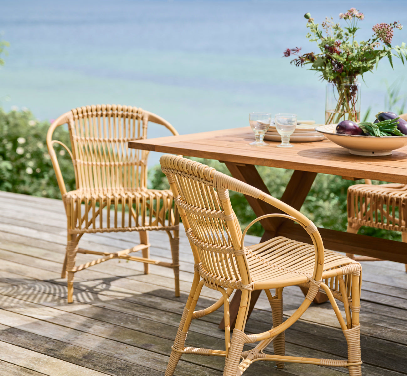 Dette er et livsstilsbillede af Robert Udendørs spisebordsstol i natural aluminium artfibre. Opstillet omkring et teakbord på et trædæk med udsigt over havet.