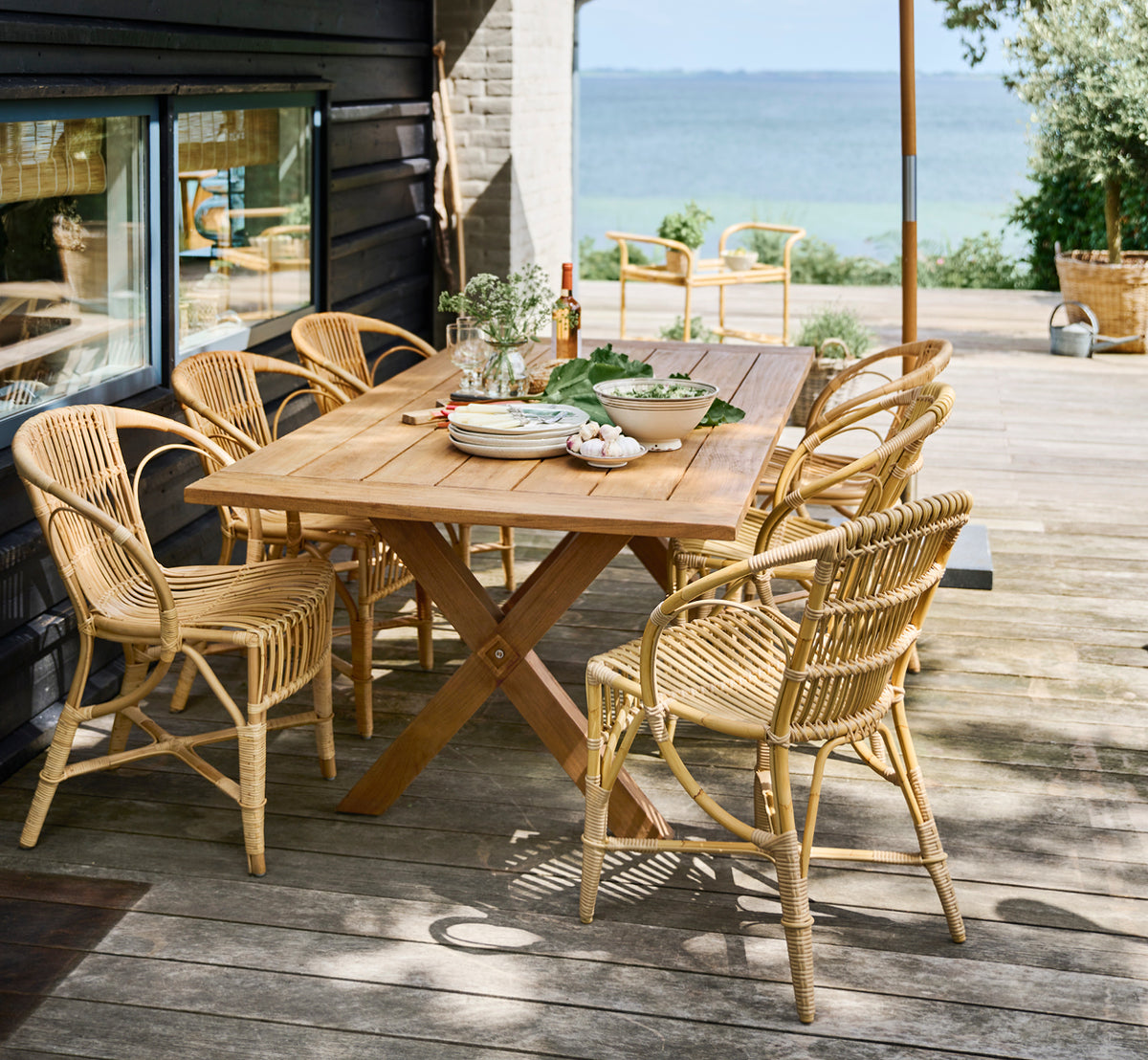Dette er et livsstilsbillede af Robert Udendørs spisebordsstol i natural aluminium artfibre. Opstillet omkring et teakbord på et trædæk med udsigt over havet.