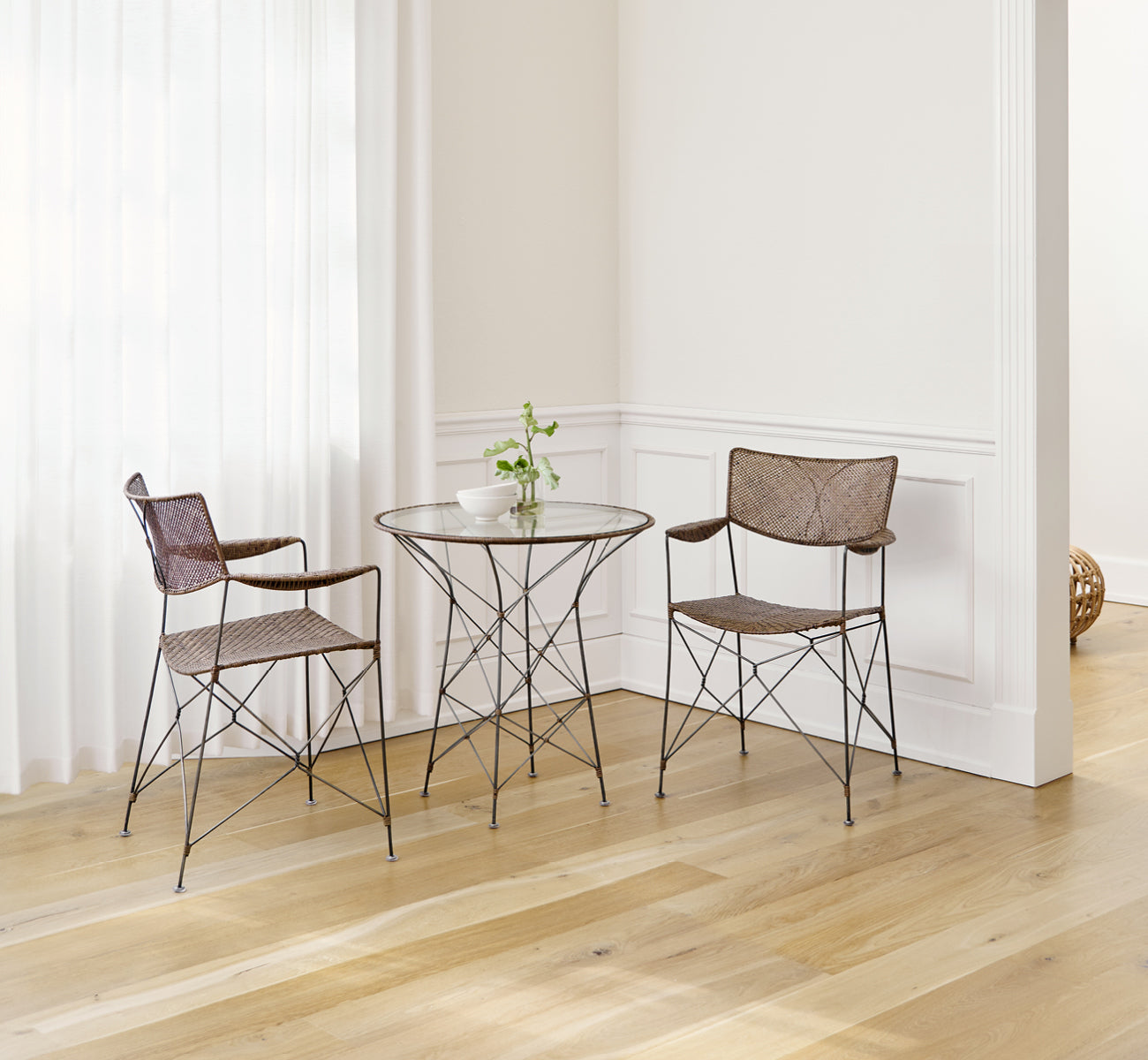 Dette er et livsstilsbillede af Rap Spisebordsstol i hazelnut rattan iron frame, i et lyst interiør med hvide panelvægge og sildebensgulv.