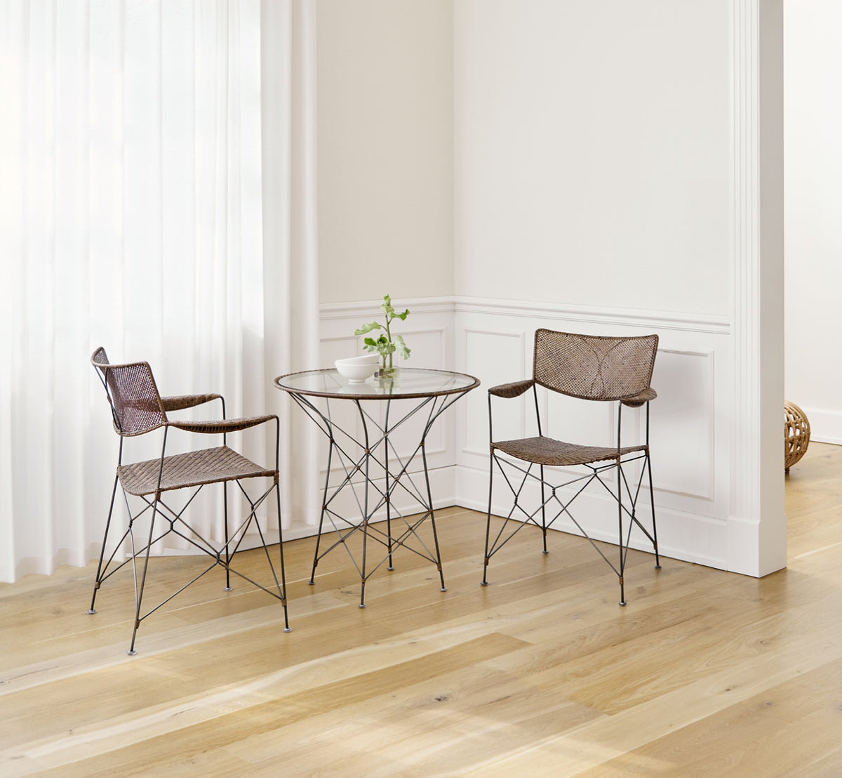 Dette er et livsstilsbillede af Rap Spisebordsstol i hazelnut rattan iron frame, i et lyst interiør med hvide panelvægge og sildebensgulv.