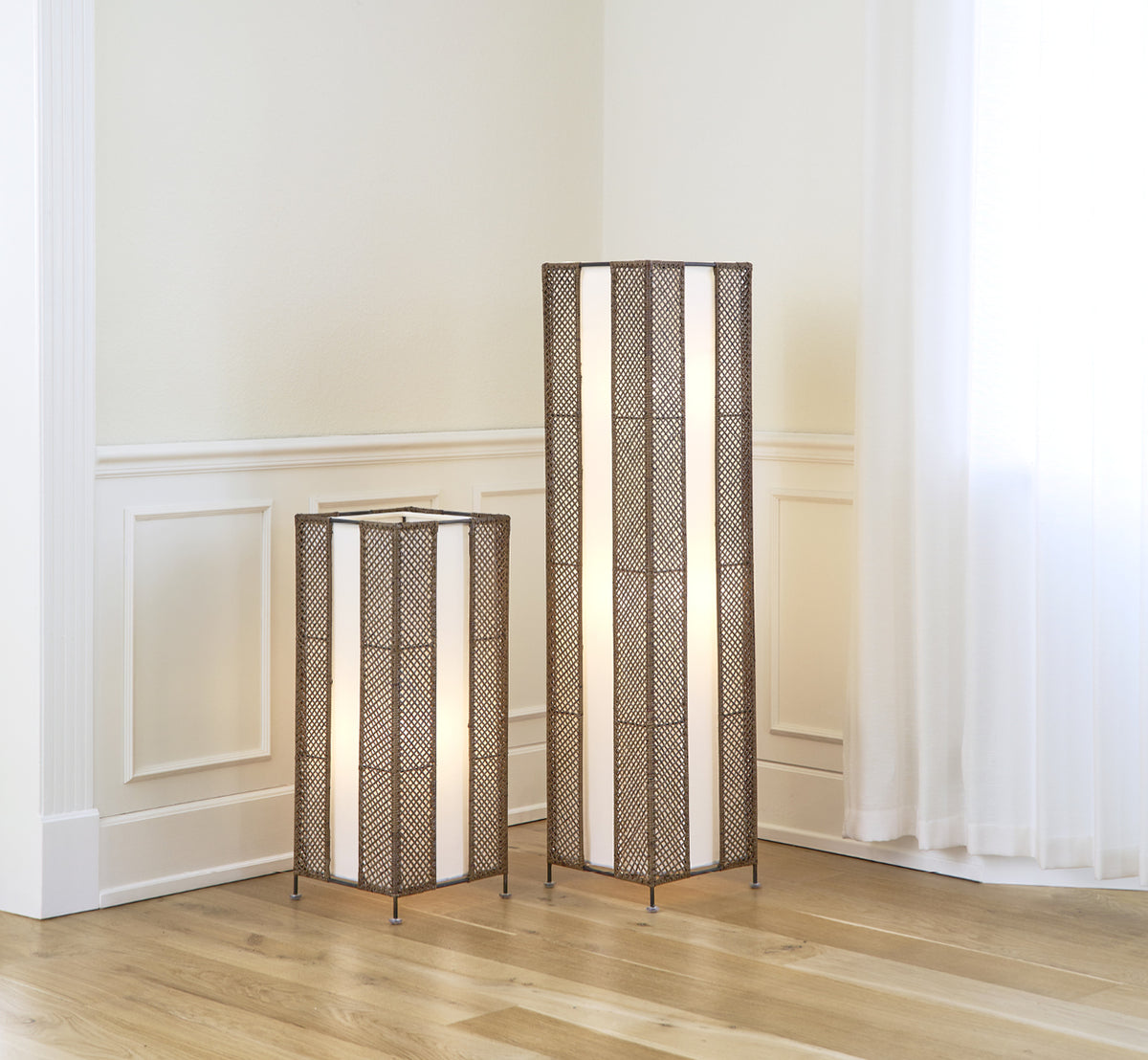Dette er et livsstilsbillede af Yamakawa Lampshade | 125 cm i hazelnut rattan iron frame. Vist som to høje gulvlamper i et lyst panelinteriør.
